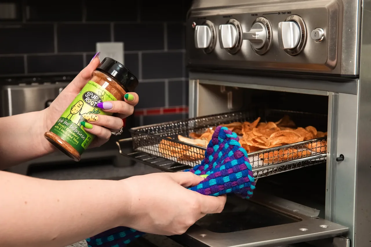 A hand holding the Herb Your Enthusiasm jar over an air fryer basket of crisp chips, while the other hand, in a blue oven mitt, pulls the basket out.