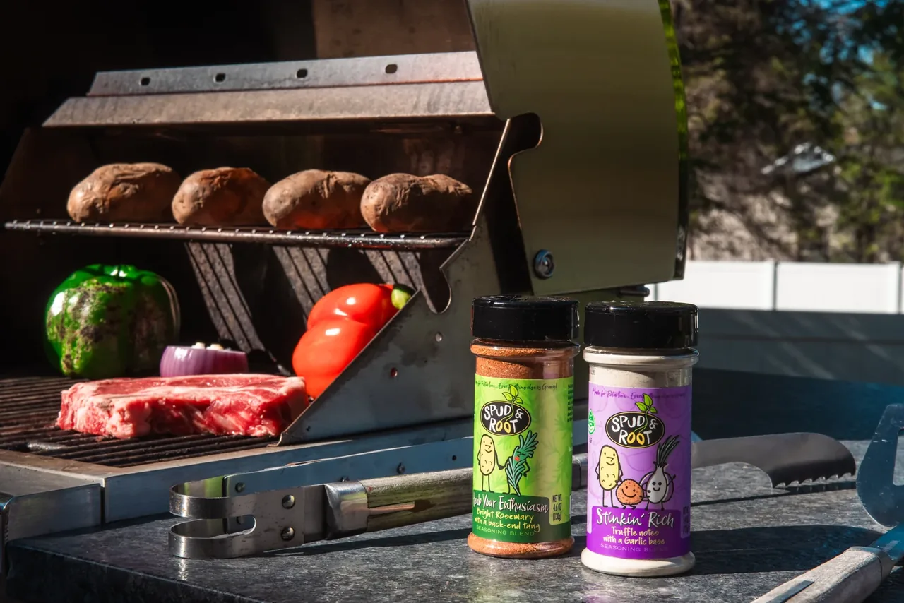 Herb Your Enthusiasm and Stinkin' Rich jars in front of an open grill holding baked potatoes, a steak and peppers.