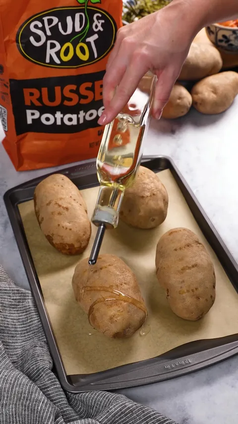 Oil being drizzled from a bottle over four russet potatoes on a baking sheet.