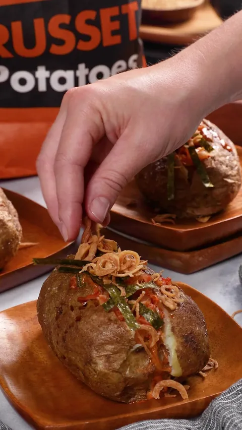 A hand sprinkling crispy fried shallots over a loaded baked potato on a wooden dish.