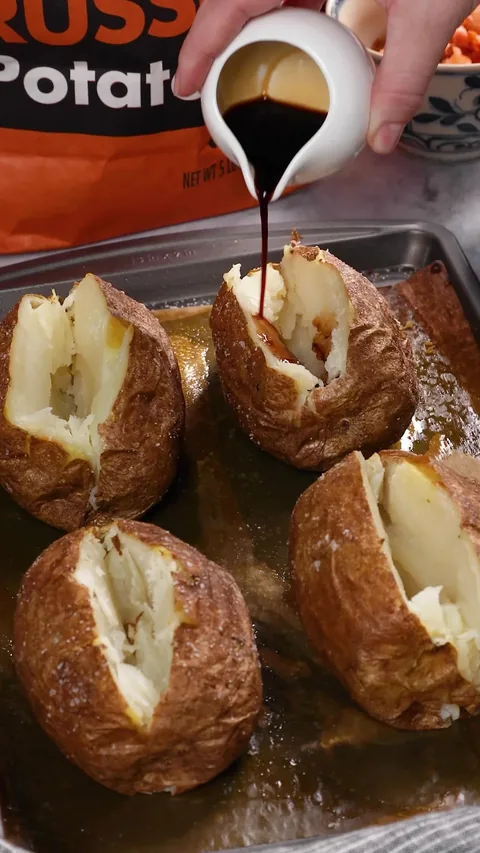 Dark soy sauce being poured from a small white cup over a split baked potato.