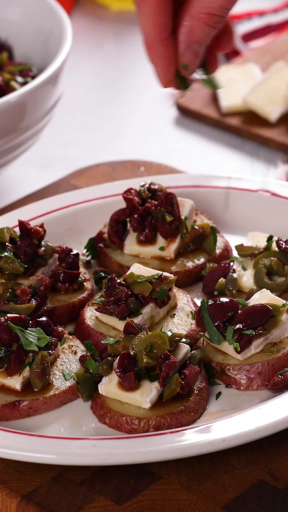 A hand sprinkling parsley over a platter of roasted red potato rounds, each topped with a slice of brie and a heap of green and kalamata olive tapenade.
