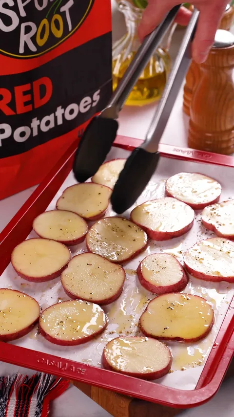 Tongs turning oiled and peppered red potato rounds on a lined red baking sheet.