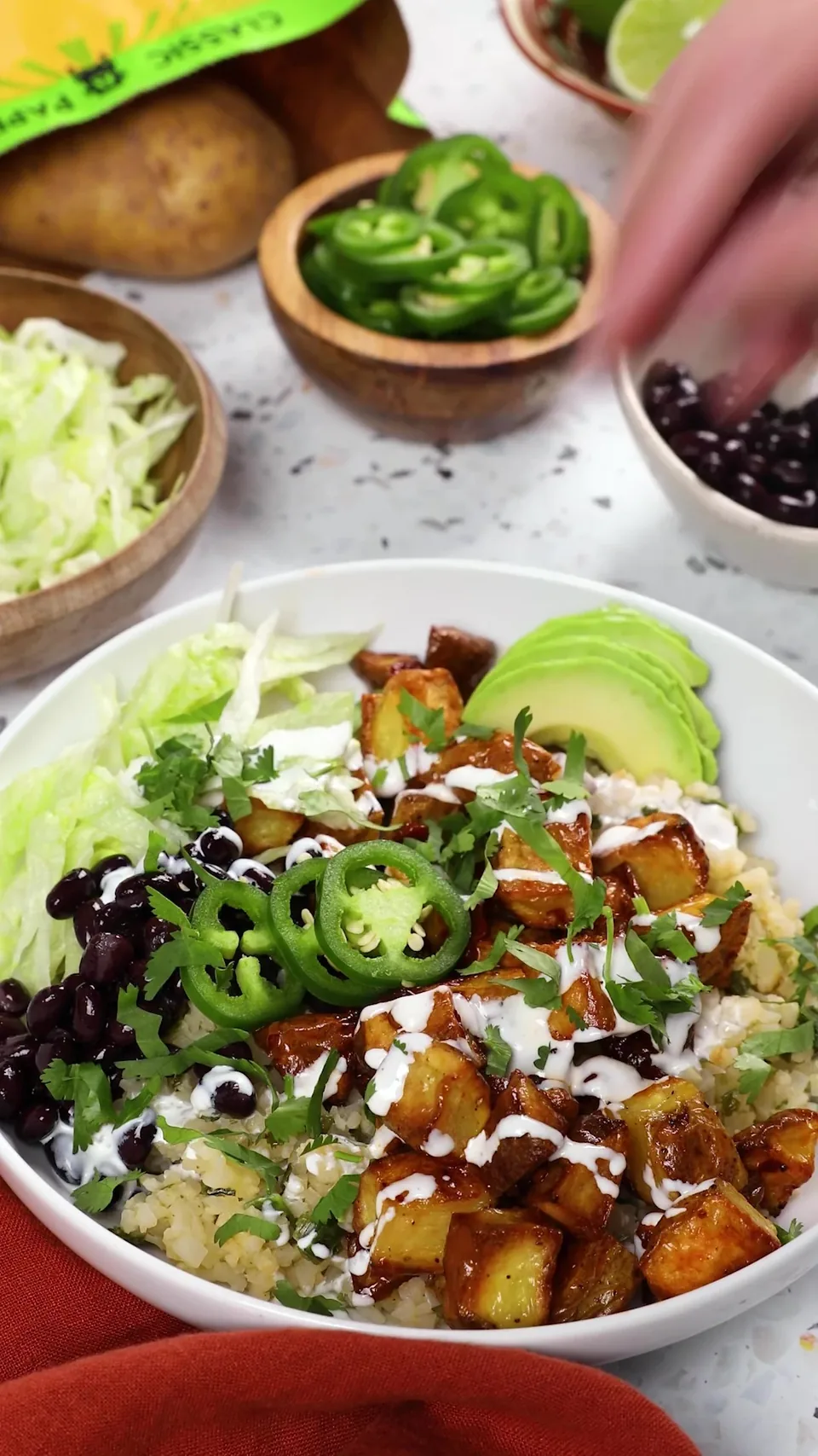 A finished chipotle potato burrito bowl with avocado, black beans, jalapeños, lettuce, crema and cilantro, surrounded by little bowls of the toppings and a few Gold potatoes.