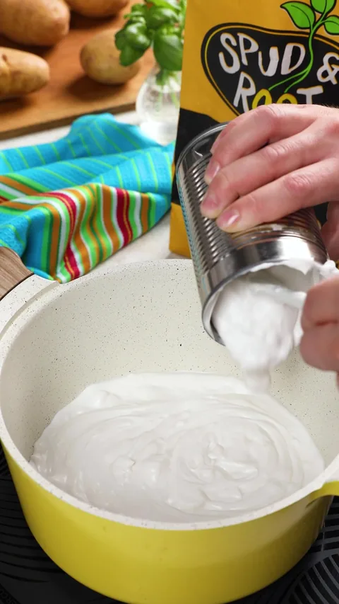 Coconut milk pouring from a can into a yellow pot, with a bag of Spud & Root Gold Potatoes behind.