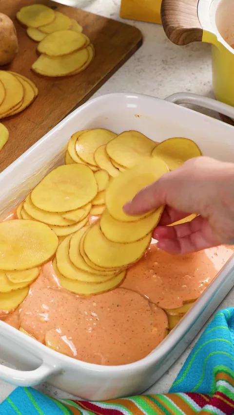 A hand laying a second layer of potato slices over the sauce.