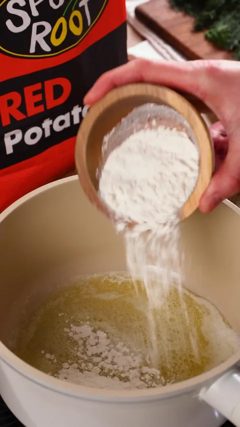 Flour pouring from a small wooden bowl into melted butter in a cream pot, with a bag of Red potatoes behind it.