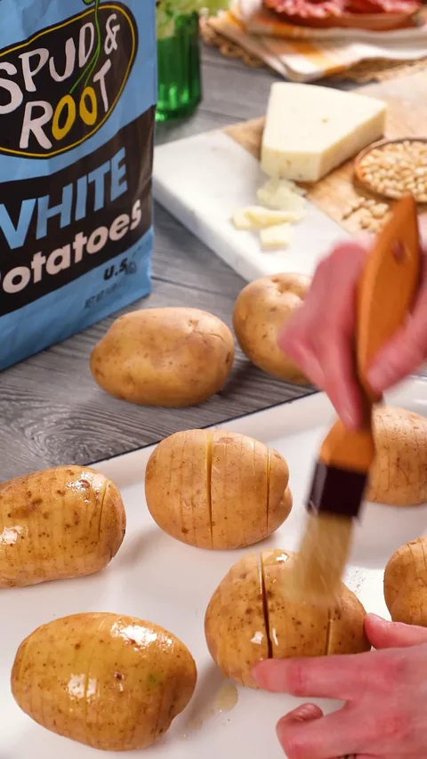 A pastry brush painting duck fat over sliced white potatoes on a white enamel tray.