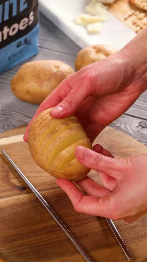 A hand holding up a white potato cut into thin slices that fan open but stay joined at the bottom.