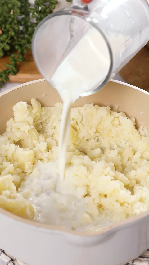 Hot milk being poured from a glass measuring cup into rough mashed potatoes.