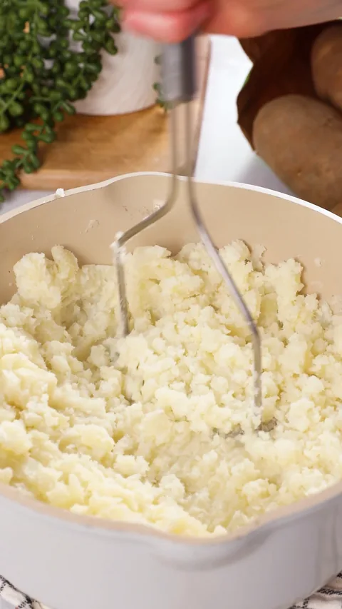 Rough, crumbly mashed potatoes in a saucepan, with the masher still in it.