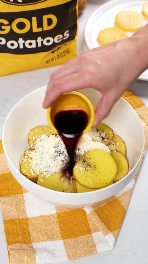 Balsamic vinegar poured from a small cup into the bowl of seasoned potato slices.