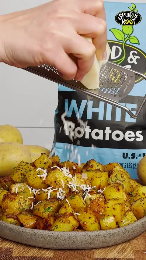 A hand grating Pecorino Romano over a plate of golden roasted potato cubes, with a bag of Spud & Root White Potatoes behind.