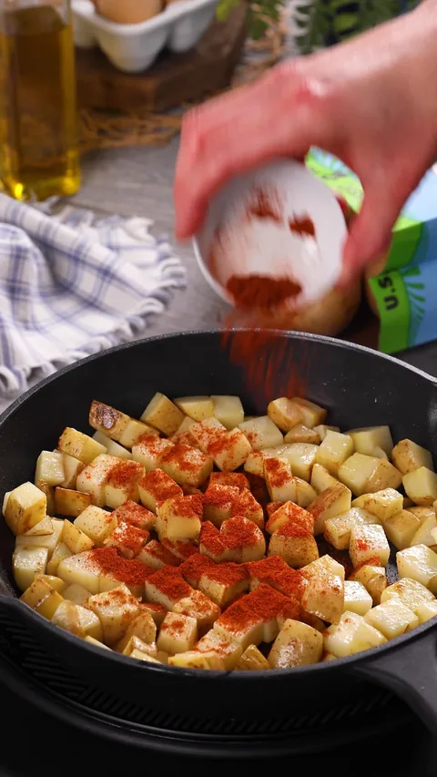 Paprika being sprinkled from a small bowl over potato cubes in a skillet.
