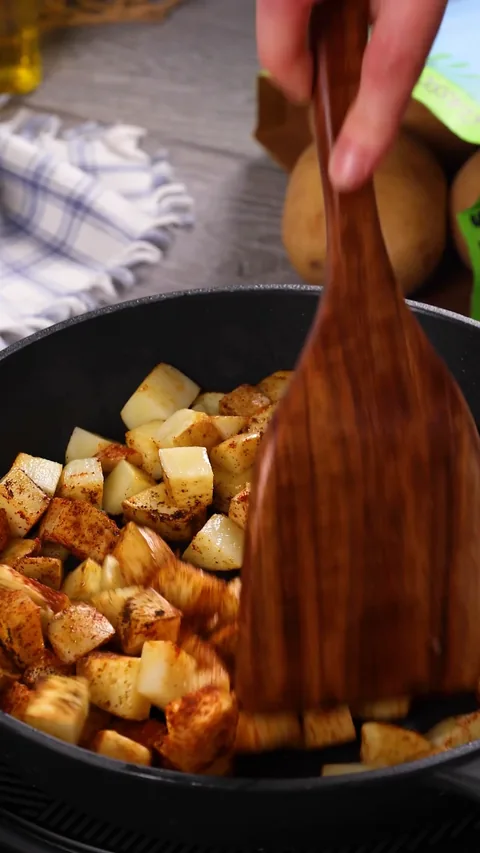 A wooden spatula turning potato cubes in a cast-iron skillet, some pale and some starting to brown.